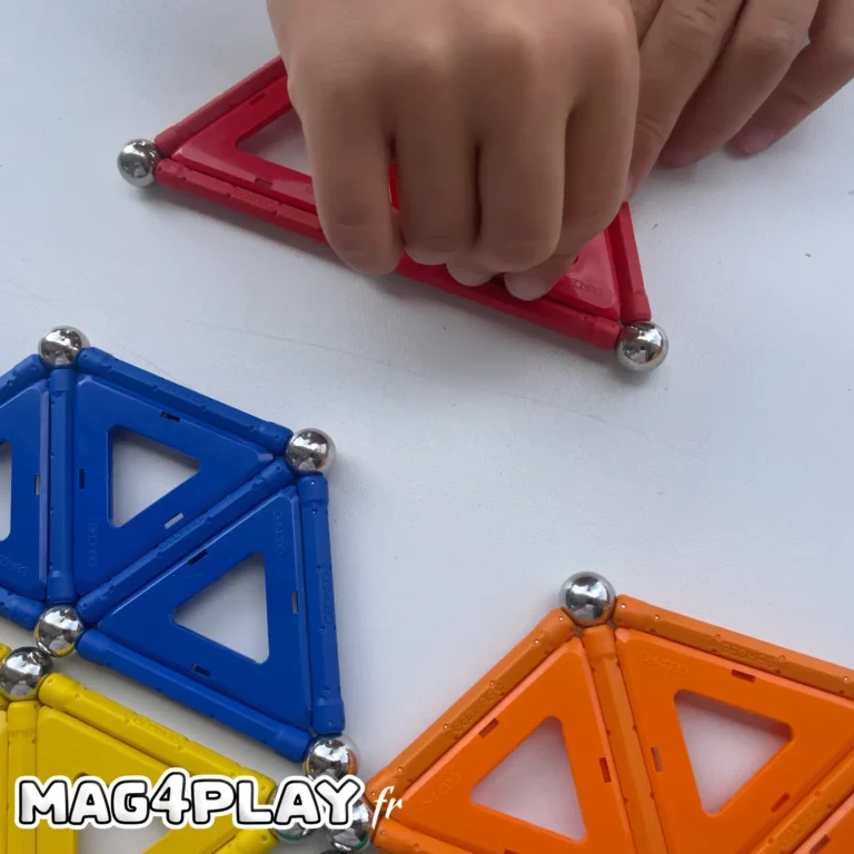 Vue rapprochée et verticale focalisée exclusivement sur les mains de deux enfants collaborant lors d'un atelier Mag4play à Lyon. Leurs doigts travaillent de concert pour connecter des barrettes Geomag à une bille d'acier centrale, créant les bases d'un pont magnétique. La lumière met en relief la texture des pièces et la concentration qui émane de ces gestes précis, capturant l'essence même de l'apprentissage par le jeu et de la coopération sans montrer les visages