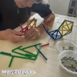 Un résident senior concentré et souriant, assemblant patiemment une structure complexe avec des barrettes et des sphères Geomag lors d’un atelier cognitif Mag4play à Lyon, dans le Rhône, en Rhône-Alpes et en Suisse romande.