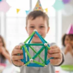 Photographie en vue subjective (POV) d'un enfant tendant vers l'objectif une structure géométrique complexe en Geomag, avec un grand sourire de fierté. Le focus est mis sur l'objet et l'expression de réussite, avec un arrière-plan flou de fête d'anniversaire Mag4play à Lyon, dans le Rhône, en région Rhône-Alpes ou en Suisse romande