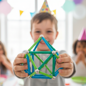 Photographie en vue subjective (POV) d'un enfant tendant vers l'objectif une structure géométrique complexe en Geomag, avec un grand sourire de fierté. Le focus est mis sur l'objet et l'expression de réussite, avec un arrière-plan flou de fête d'anniversaire Mag4play à Lyon, dans le Rhône, en région Rhône-Alpes ou en Suisse romande